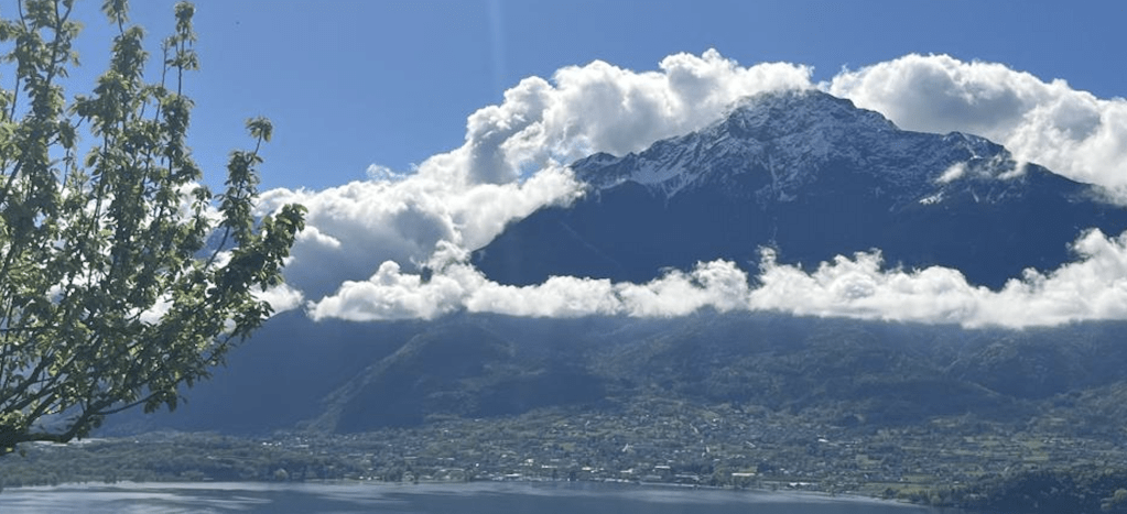 A panoramic view of a mountain range with snow-capped peaks under a bright blue sky, surrounded by fluffy white clouds. In the foreground, a tree is partially visible, while a town and a lake can be seen below the mountains.